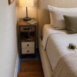 Narrow charging nightstand fitted between an upholstered bed and wall with its footprint marked for measurement