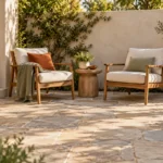 AI-created compact courtyard patio with two wood armchairs, cream cushions, sage and rust accents, a small round table, a planter at the edge and open floor space in front; illustrative furniture.