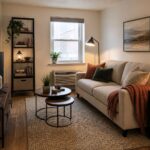 Warm small industrial living room with a neutral sofa, nesting wood tables, narrow black metal shelf, glass lamps, a textured rug, and a clear walking path